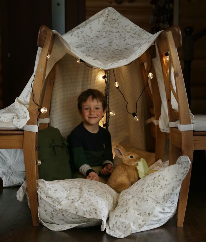 Niño en casita hecha con cobijas, almohadas y sillas.