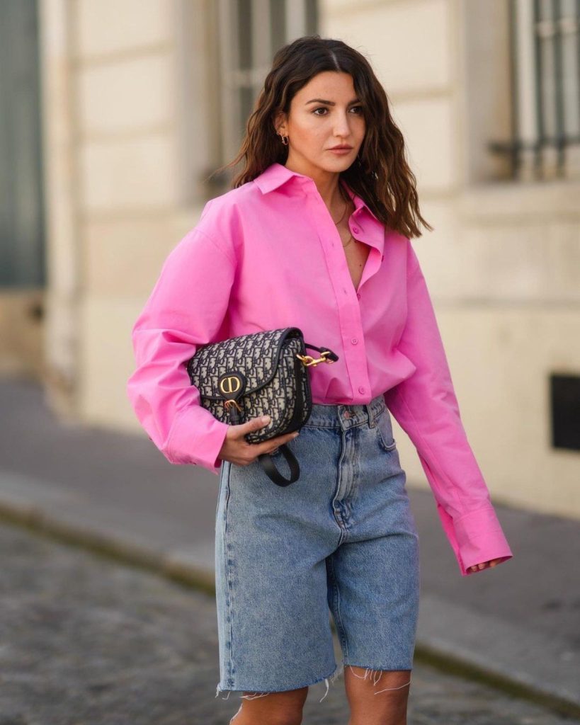 Imagen de mujer caminando. Ella viste una camisa over size color rosa y pantalones cortos de jean, en su mano lleva un bolso de color negro con detalles en blanco y dorado.