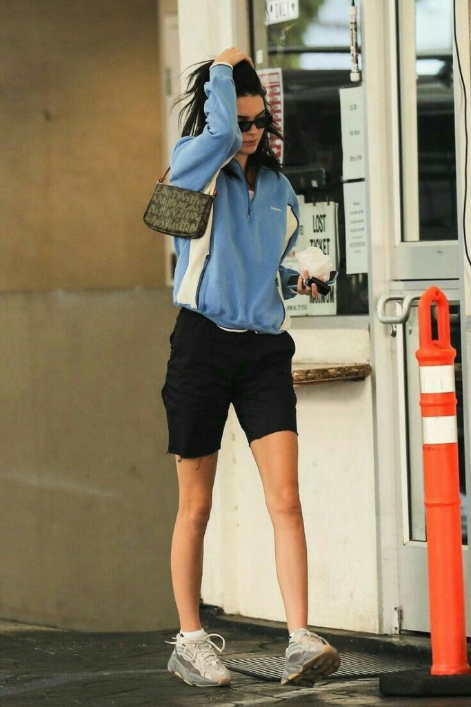 Fotografía de Kendall Jenner caminando por la calle.  La modelo está usando un pullover con cierre en el cuello de color celeste con dos franjas blancas a los costados, pantalones cortos de color negro, zapatillas de correr blancas.
Los accesorios que lleva son una baguette bag de color gris y unos lentes de sol. 