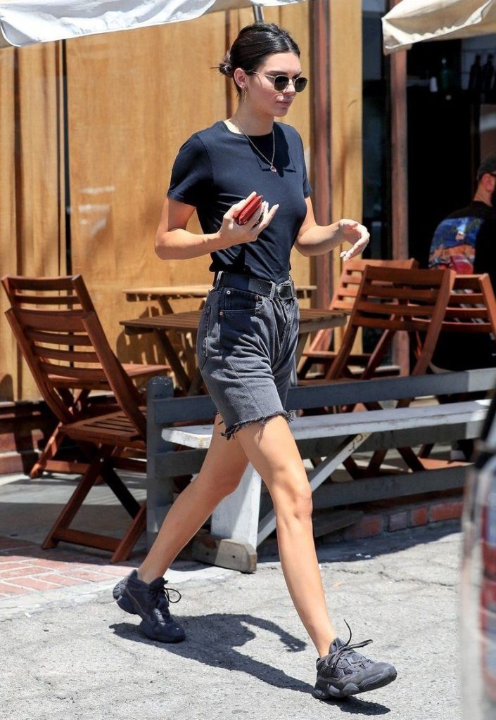 Imagen de Kendall Jenner caminando por la calle. Lleva puesto una remera básica negra, dad shorts negros , zapatillas de correr negras. Lentes de sol y cinto también de color negro.