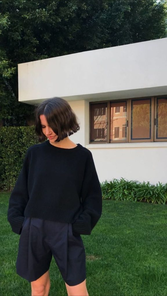 Imagen de una chica frente a una fachada de una casa, en el jardín mirando hacia abajo, viste un Sweater tejido negro y unos pantalones cortos hasta la rodillas anchos, de color negro. 