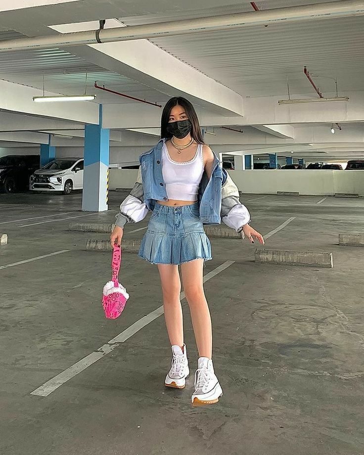 Imagen de una joven en un estacionamiento subterráneo. Lleva puesta una  remera de tirantes color blanco con una campera de jean , tiene una falda de jean, un bolso de mano color rosa y unas converse de suela track blanca.