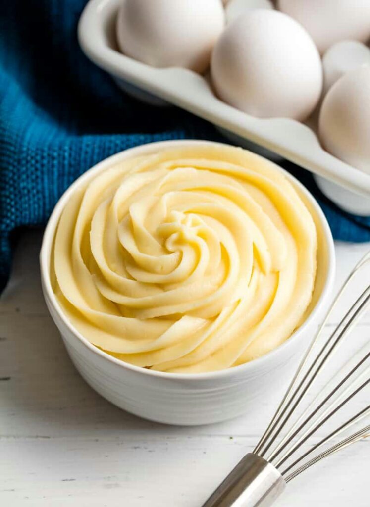 Crema pastelera colocada con manga pastelera con boquilla risada en un bowl de cerámica blanca, por detrás se ve un mantel azul turquesa, un batidor de mano de acero y media docena de huevos.